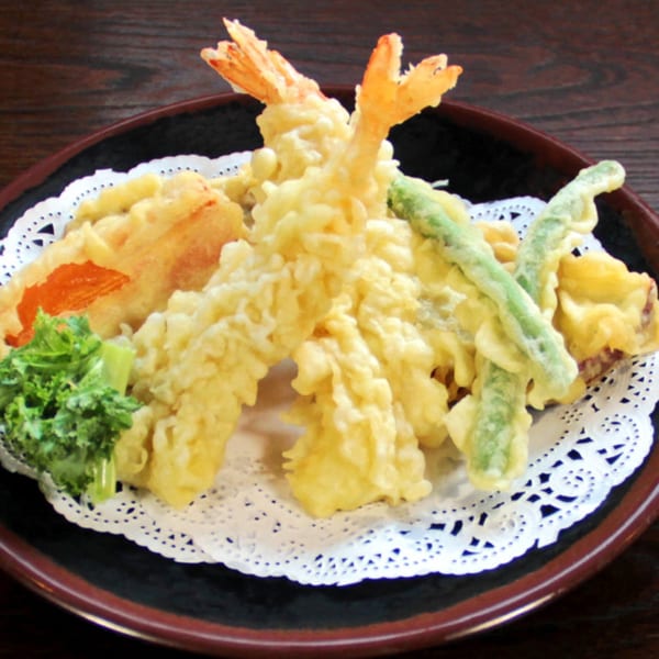 A black plate with a white doily holds golden-fried tempura shrimp, green beans, and vegetables.